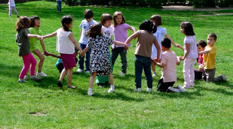 Niños de distintas edades cogidos de la mano formando un corro sobre el césped de un parque.
